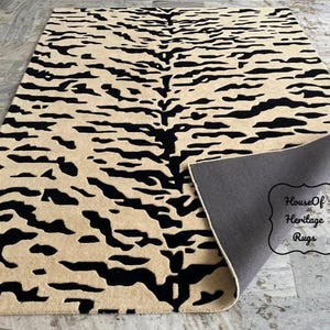 May include: A rectangular area rug featuring a black tiger stripe pattern on a beige background. The rug is partially folded at one corner, revealing a solid dark gray backing. A decorative label reads House Of Heritage Rugs.