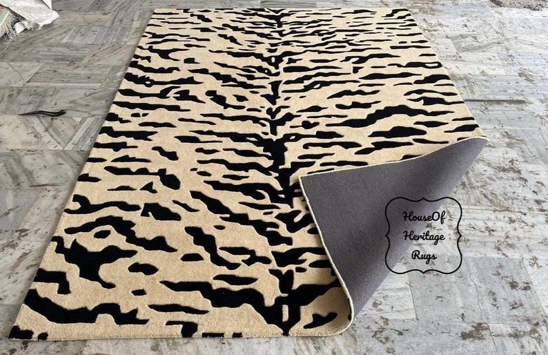 May include: A rectangular area rug featuring a black tiger stripe pattern on a beige background. The rug is partially folded at one corner, revealing a solid dark gray backing. A decorative label reads House Of Heritage Rugs.