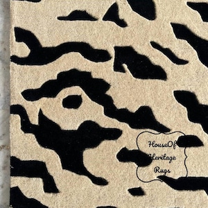 May include: A rectangular rug featuring a high-contrast, abstract pattern of black organic shapes set against a beige background. The surface has a textured, tufted appearance. A black outlined label with the text House Of Heritage Rugs is visible.