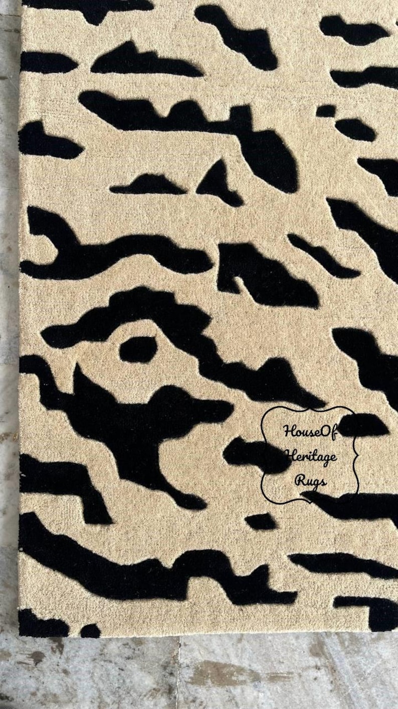 May include: A rectangular rug featuring a high-contrast, abstract pattern of black organic shapes set against a beige background. The surface has a textured, tufted appearance. A black outlined label with the text House Of Heritage Rugs is visible.