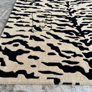 May include: A rectangular area rug featuring a high-contrast black and cream abstract pattern with organic, flowing shapes. The rug has a textured, tufted surface and includes a central logo stamp that reads House of Heritage Rugs.