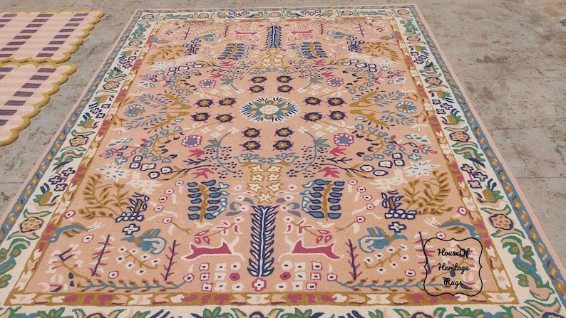 Peut inclure: Un grand tapis rectangulaire avec un fond p&ecirc;che et des motifs floraux et animaliers complexes dans des tons de bleu, vert, rose et or. Le tapis a une bordure d&eacute;taill&eacute;e et un motif de m&eacute;daillon central. Un plus petit tapis ray&eacute; est visible.