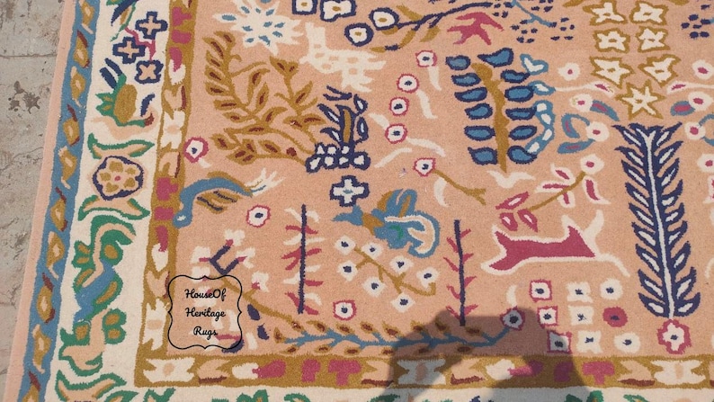 Peut inclure: Gros plan sur un tapis p&ecirc;che avec un motif floral et faunique d&eacute;taill&eacute;. Le tapis pr&eacute;sente une bordure avec des accents bleus, verts et cr&egrave;me. Le motif central comprend des fleurs, des feuilles et des figures d'animaux stylis&eacute;es.