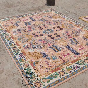 Peut inclure: Un grand tapis rectangulaire avec un motif floral et g&eacute;om&eacute;trique d&eacute;taill&eacute; dans des tons de p&ecirc;che, de bleu, de vert et de cr&egrave;me. Le tapis a une bordure avec des motifs floraux et est pos&eacute; sur une surface en b&eacute;ton. Un plus petit tapis &agrave; motifs &agrave; carreaux est &eacute;galement visible.