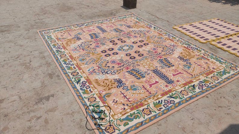 Peut inclure: Un grand tapis rectangulaire avec un motif floral et g&eacute;om&eacute;trique d&eacute;taill&eacute; dans des tons de p&ecirc;che, de bleu, de vert et de cr&egrave;me. Le tapis a une bordure avec des motifs floraux et est pos&eacute; sur une surface en b&eacute;ton. Un plus petit tapis &agrave; motifs &agrave; carreaux est &eacute;galement visible.