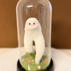 May include: A whimsical white plush figure with a smiling face and two legs, displayed under a clear glass dome. The figure stands on a bed of green moss with small pink flowers, set on a black base.