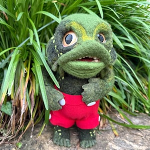 May include: A handmade felt creature with a frog-like face, green body, and red shorts. The creature has large, expressive eyes, a wide mouth, and small arms. The shorts have white buttons. The creature is standing in front of green foliage.