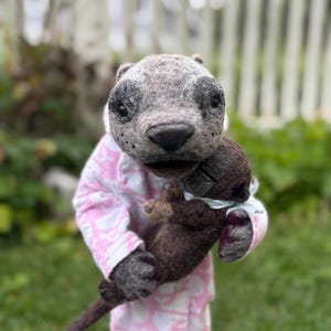 May include: A handmade felt otter doll with a smaller otter doll. The larger otter has gray fur with black accents around the eyes and nose. The doll is wearing a pink and white patterned onesie. The smaller otter is brown.