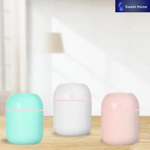 Op de afbeelding: Drie kleine, ronde, ultrasone luchtbevochtigers. De luchtbevochtigers zijn wit, lichtblauw en lichtroze. Ze hebben een kleine opening aan de bovenkant om water en etherische oliën toe te voegen.