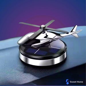 Puede incluir: Un ambientador para coche con forma de helicóptero plateado y negro. La aeronave en miniatura cuenta con una base de panel solar y aspas giratorias. El producto está diseñado para colocarse en el salpicadero del coche.