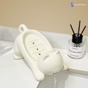 Op de afbeelding: Een witte keramische katvormige zeepbakje met een sleufvormig oppervlak en een staart. De kop van de kat heeft een tuit en er stroomt water uit. Naast het zeepbakje staat een kleine glazen fles met zwarte rietjes en de tekst "Paris Rain Alley".