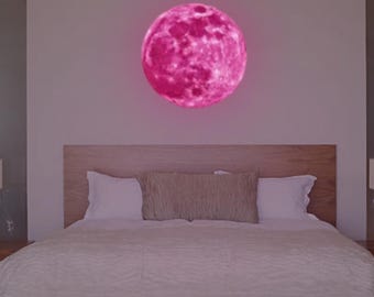 May include: A pink glow-in-the-dark moon wall decal, featuring a detailed moon surface, is displayed above a white bed with a wooden headboard.