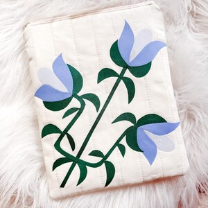 May include: Cream-colored quilted pouch with a floral design. The design features three stylized blue and white flowers with green stems and leaves. The pouch has a rectangular shape and a simple, minimalist aesthetic.