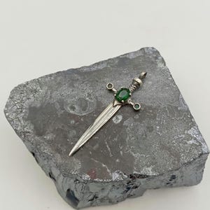 May include: A silver sword pendant with a green gemstone set in the center. The sword has a detailed design and is accented with smaller green gemstones.