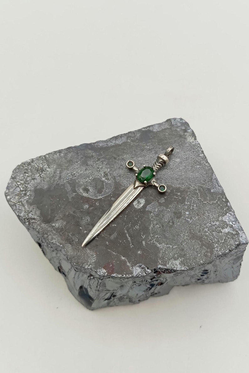 May include: A silver sword pendant featuring a green gemstone set in the centre. The sword has an intricate design and is accented with smaller green gemstones.