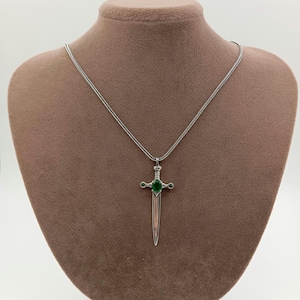 May include: A silver-colored pendant necklace featuring a sword-shaped charm. The charm has a green gemstone at the center and smaller green accents. The necklace is displayed on a brown mannequin.