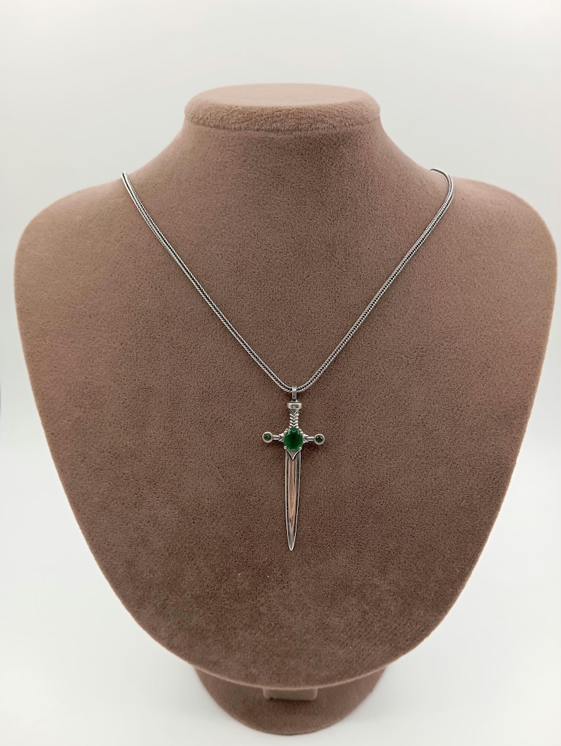 May include: A silver-colored pendant necklace featuring a sword-shaped charm. The charm has a green gemstone at the center and smaller green accents. The necklace is displayed on a brown mannequin.