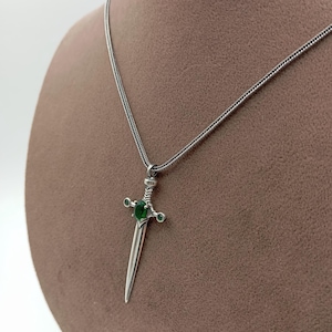 May include: A silver-toned pendant necklace featuring a dagger design. The dagger has a green gemstone in the center and smaller green stones on the crossguard. The necklace is displayed on a brown mannequin.