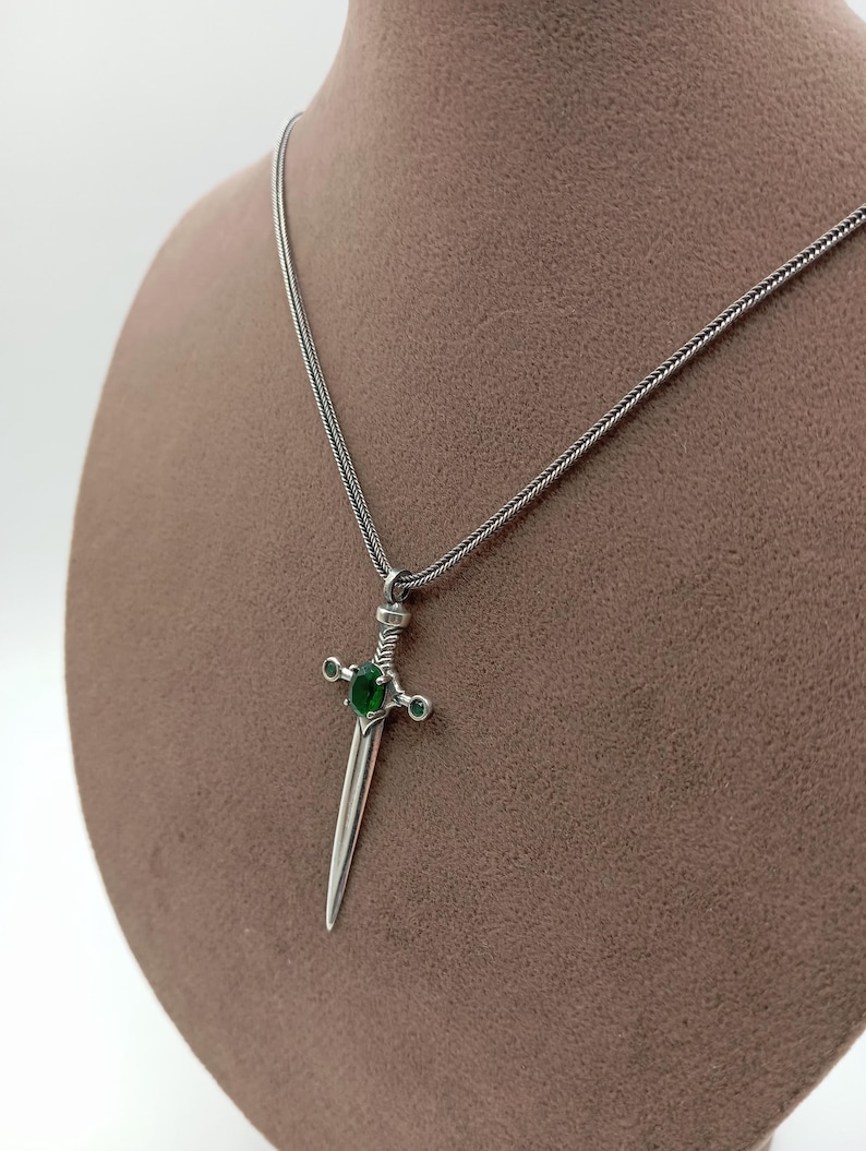 May include: A silver-toned pendant necklace featuring a dagger design. The dagger has a green gemstone in the center and smaller green stones on the crossguard. The necklace is displayed on a brown mannequin.