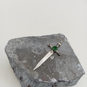 May include: A silver sword pendant with a green gemstone set in the hilt. The pendant is resting on a grey rock.