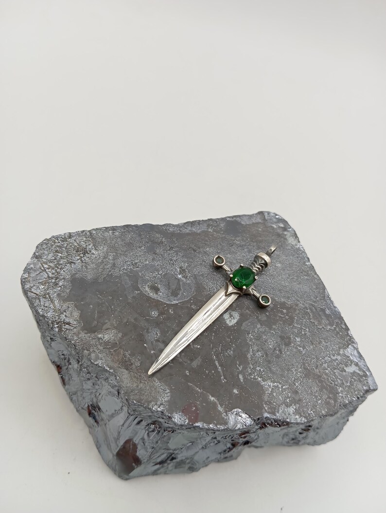 May include: A silver sword pendant with a green gemstone set in the hilt. The pendant is resting on a grey rock.