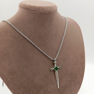 May include: A silver-colored pendant necklace featuring a sword-shaped charm. The charm has a green gemstone at the center and smaller green gems on the crossguard. The necklace is displayed on a brown mannequin.