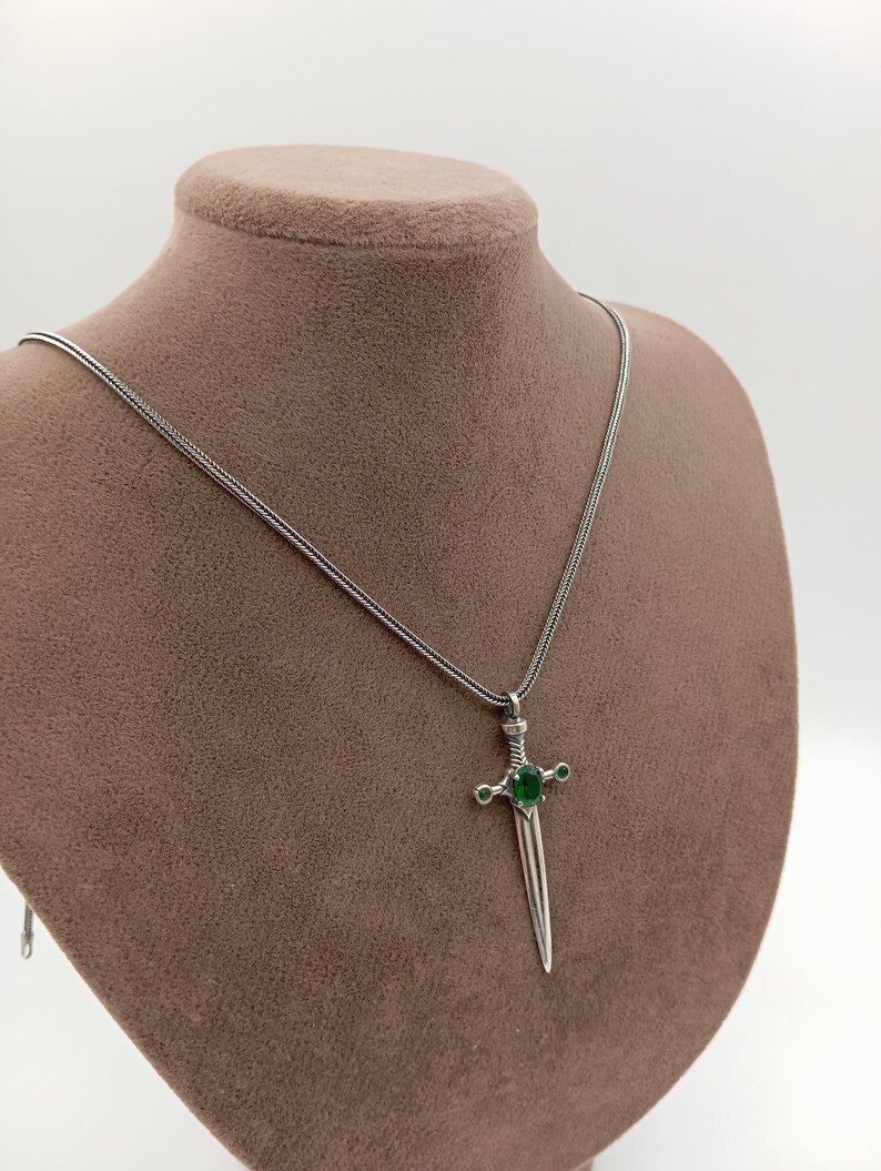 May include: A silver-colored pendant necklace featuring a sword-shaped charm. The charm has a green gemstone at the center and smaller green gems on the crossguard. The necklace is displayed on a brown mannequin.