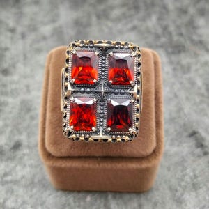 May include: A rectangular silver ring featuring four large, vibrant red gemstones. The ring has a detailed, ornate design with gold-colored accents and a dark border. The ring is displayed on a brown velvet surface.