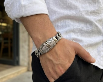 May include: A heavy silver-tone chain bracelet featuring a central rectangular plaque with intricate engraved scrollwork patterns and gold-tone accents, connected to thick, interlocking curb-style links that wrap around the wrist.