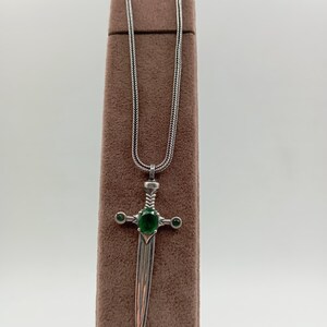 May include: A silver pendant necklace featuring a sword design. The sword has a green gemstone in the center of the hilt and two smaller green gemstones on either side. The necklace chain is a fine, textured silver.