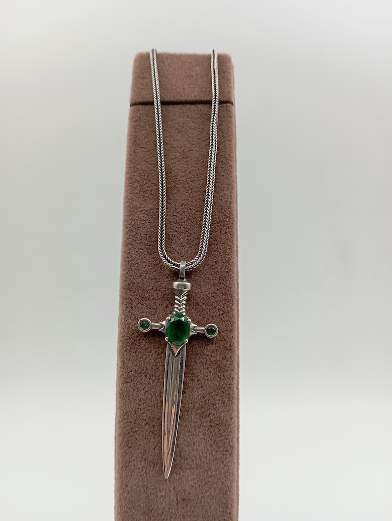 May include: A silver pendant necklace featuring a sword design. The sword has a green gemstone in the center of the hilt and two smaller green gemstones on either side. The necklace chain is a fine, textured silver.