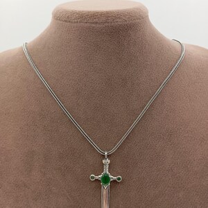 May include: A silver-toned pendant necklace featuring a sword design with a green gemstone at the center and two smaller green gemstones on either side. The sword has a detailed hilt and a polished blade, suspended from a delicate chain.