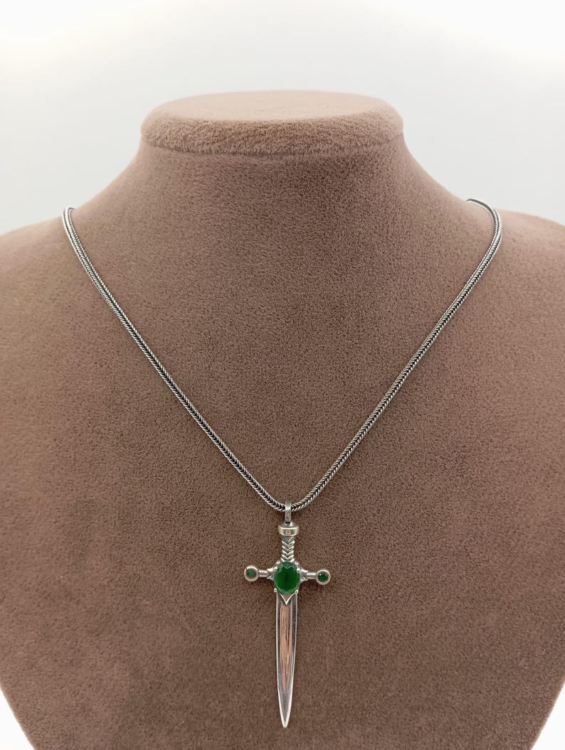 May include: A silver-toned pendant necklace featuring a sword design with a green gemstone at the center and two smaller green gemstones on either side. The sword has a detailed hilt and a polished blade, suspended from a delicate chain.