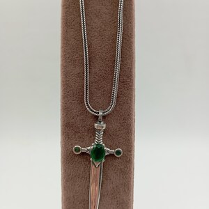 May include: Silver-toned pendant necklace featuring a dagger design. The dagger has a green gemstone at the center and two smaller green gemstones on the crossguard. The blade has a copper-colored finish.
