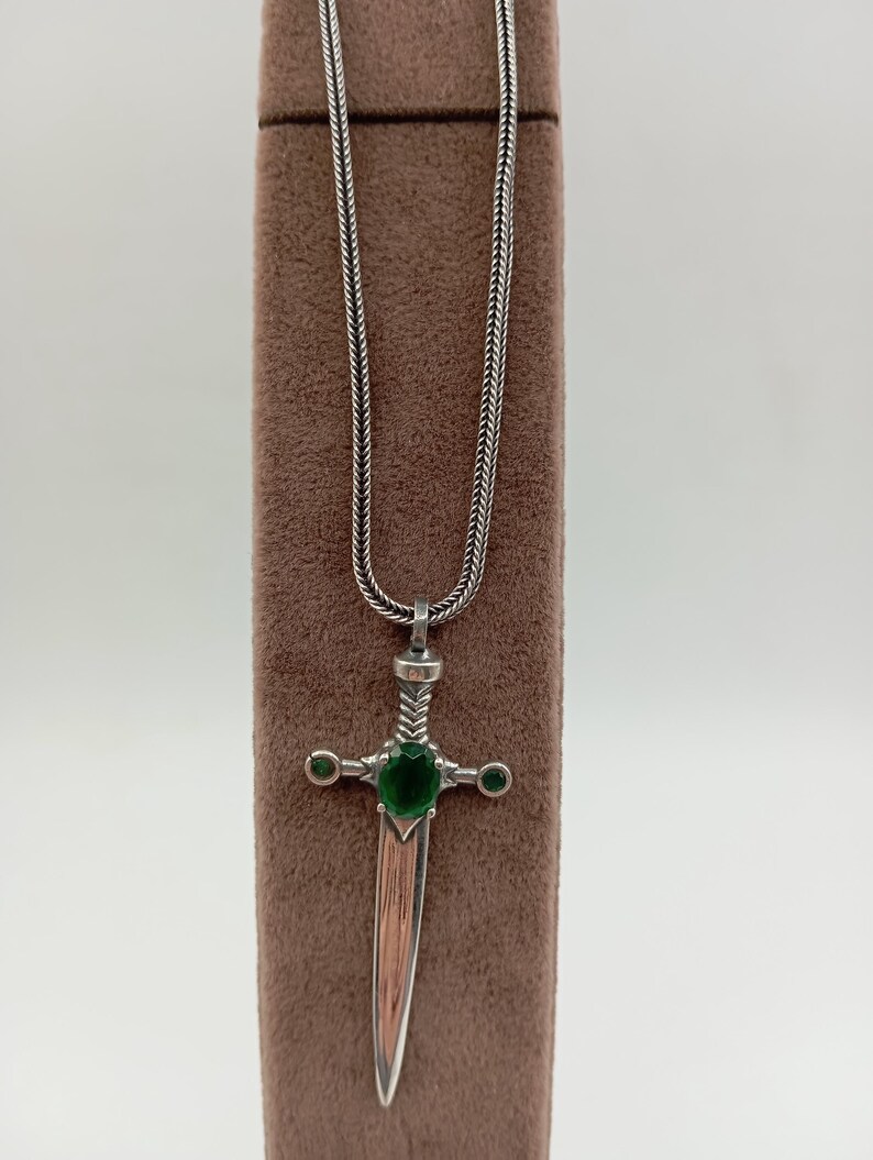 May include: Silver-toned pendant necklace featuring a dagger design. The dagger has a green gemstone at the center and two smaller green gemstones on the crossguard. The blade has a copper-colored finish.