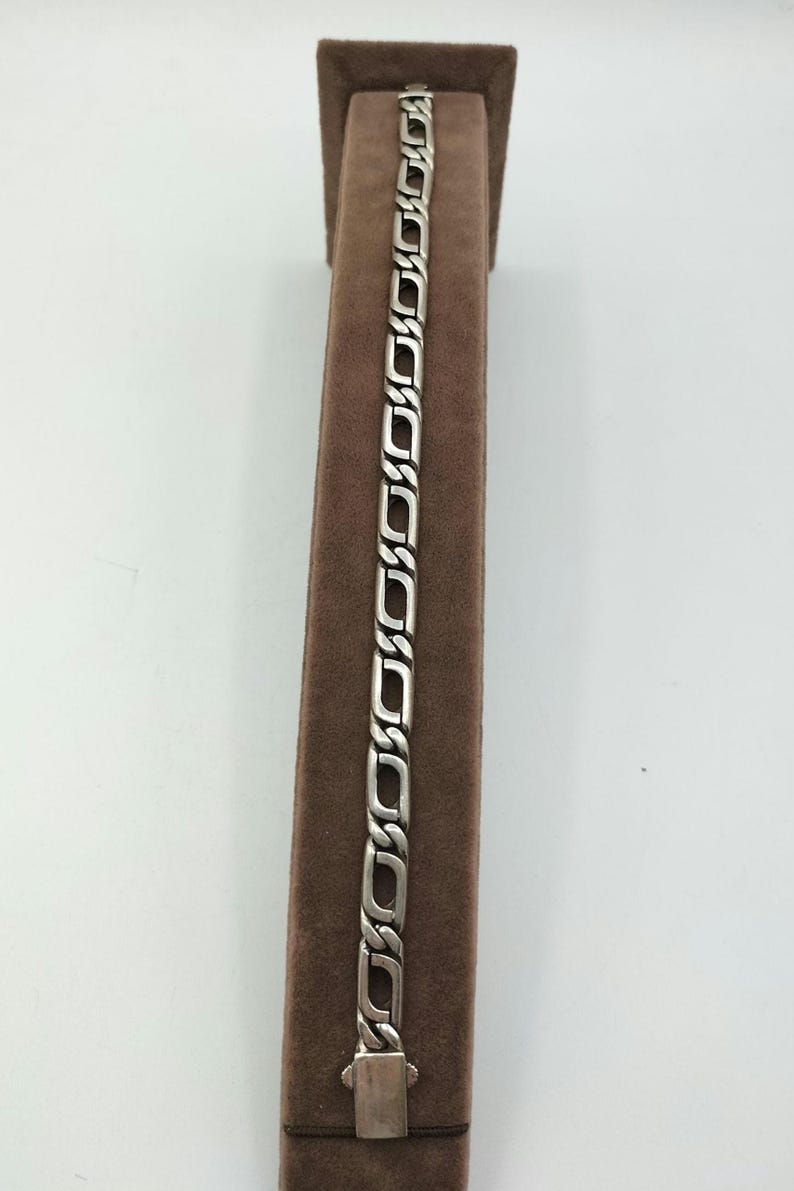 May include: A silver chain bracelet with a box clasp. The bracelet is displayed on a brown velvet stand.