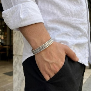 Pode incluir: Uma pulseira de prata com um padrão trançado central ladeado por duas linhas horizontais finas e lisas. O metal tem um acabamento polido com áreas rebaixadas escurecidas que destacam a textura tecida intrincada do design.