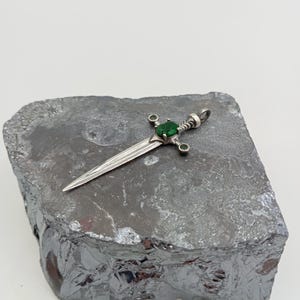 May include: A silver sword pendant with a green gemstone set in the hilt. The pendant is resting on a piece of grey rock.