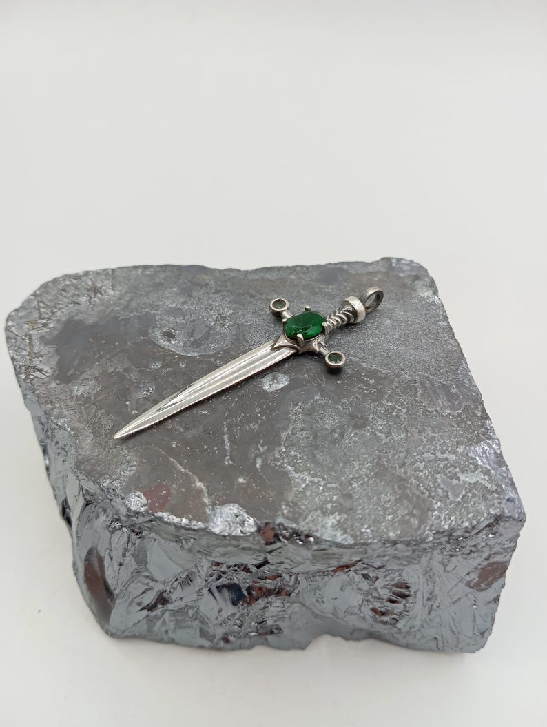 May include: A silver sword pendant with a green gemstone set in the hilt. The pendant is resting on a piece of grey rock.
