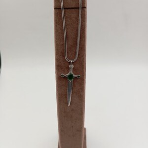 May include: A silver-toned pendant necklace featuring a sword design with a green gemstone at the center. The sword pendant is suspended from a delicate chain. The necklace is displayed on a brown jewelry stand against a white background.