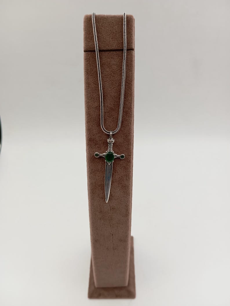 May include: A silver-toned pendant necklace featuring a sword design with a green gemstone at the center. The sword pendant is suspended from a delicate chain. The necklace is displayed on a brown jewelry stand against a white background.