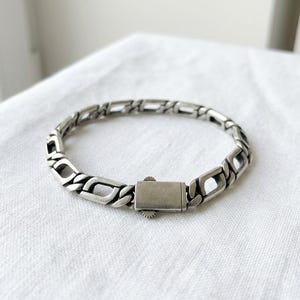 May include: A silver-toned metal chain bracelet featuring rectangular, open-link segments connected by smaller, rounded links. The bracelet includes a rectangular box clasp closure with a small, serrated edge detail on the side.