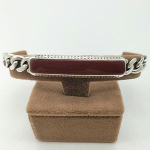 Puede incluir: Una pulsera de plata con una incrustación de piedra roja. La pulsera está en una caja de exposición de terciopelo marrón.