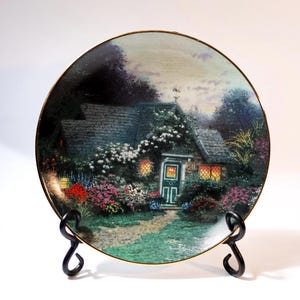 May include: A round porcelain plate with a gold rim features a painting of a cottage with a green door and a garden with flowers. The plate is on a black metal stand.