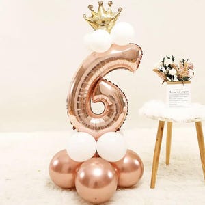 May include: A rose gold number six balloon with a gold crown on top. The balloon is surrounded by white and rose gold balloons.