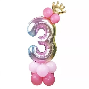 May include: A rainbow iridescent number 3 balloon with a gold crown on top. The balloon is surrounded by pink and white balloons.