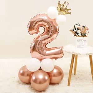 May include: Rose gold number two balloon with a crown on top. The balloon is surrounded by smaller rose gold and white balloons.