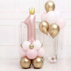 May include: A pink number one balloon topped with a gold crown, with a cluster of gold, white, and pink balloons. Ideal for a first birthday party, the balloons are arranged in a decorative display.