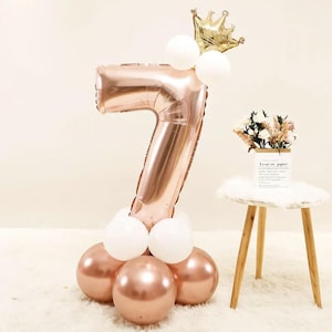 May include: Rose gold number seven balloon with a gold crown on top. The balloon is surrounded by white and rose gold balloons.