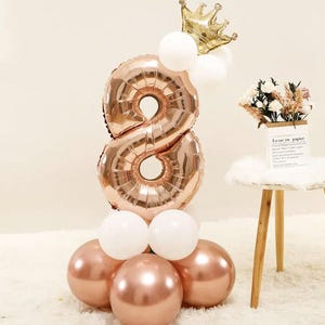 May include: A gold crown sits atop a rose gold number eight balloon. The balloon is surrounded by white and rose gold balloons. The balloons are arranged in a tower shape. The background is a white surface.